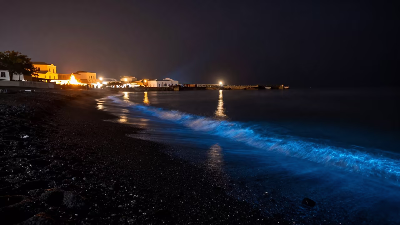 Bioluminescent Waves on Greek Black Sand Beach in beside a lantern-dotted harbor in Greece