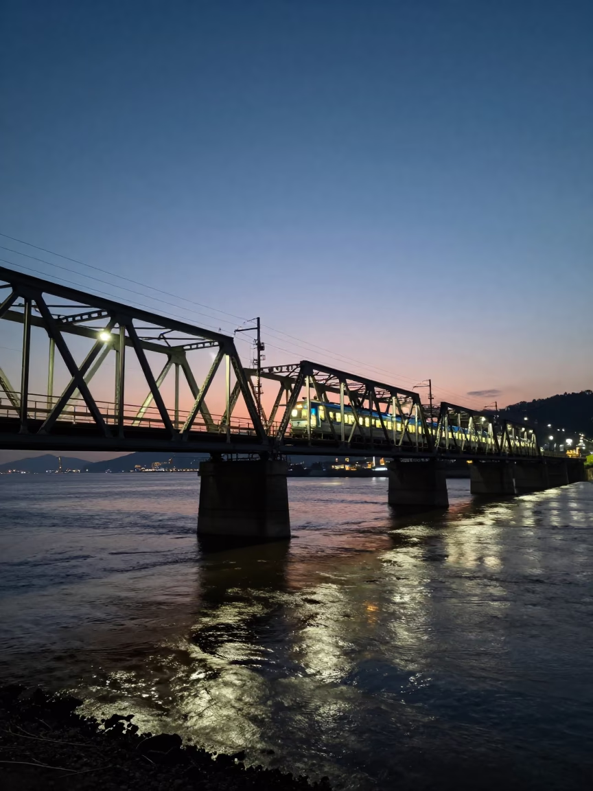 Night train crossing floodlit iron bridge in Busan before sunrise with coastal city lights in in Busan, South Korea